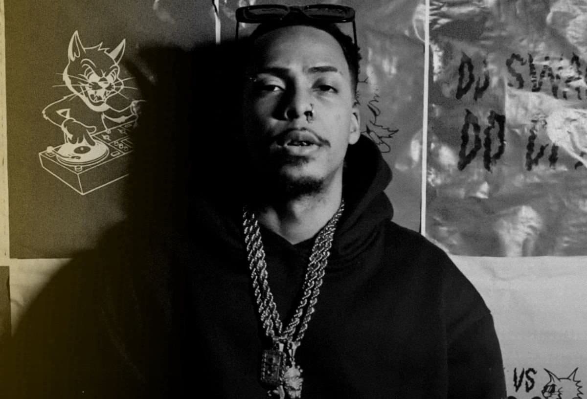 Swag do Complexo in black and white, with a chain around his neck, in front of a wall covered in posters