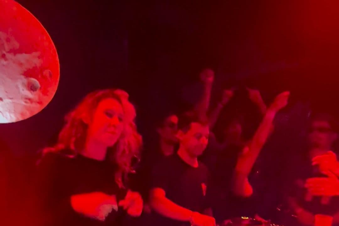 Two people behind the CDJs, with the Moon Club moon behind them and the crowd's arms in the air under red light